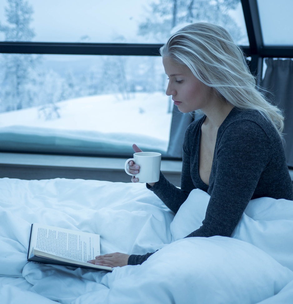 Blond model girl reading in bed with a coffee in the igloo with snow outside